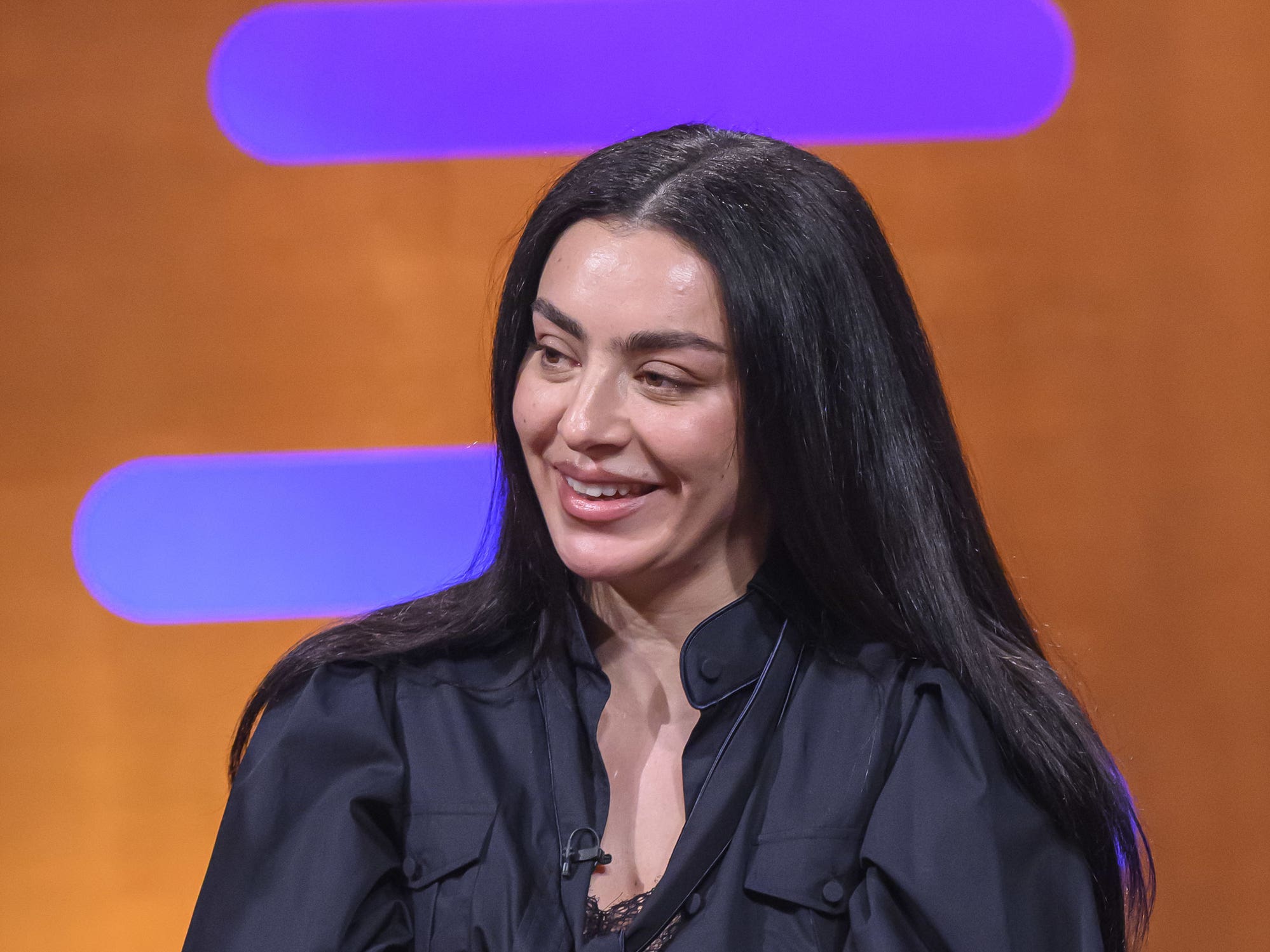 Charli XCX in a leather jacket smiling while sitting on a red couch